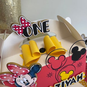 May include: A red and white Minnie Mouse themed party favor box with a yellow bow, a black and white Minnie Mouse head, and the words "ONE" and "AZIYAH" on the front.