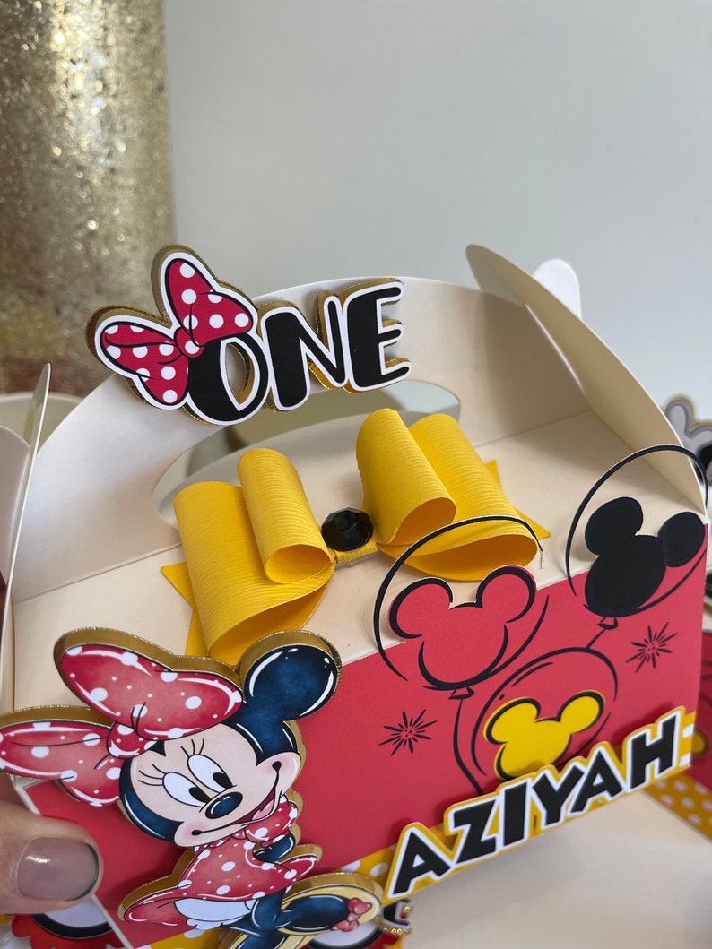 May include: A red and white Minnie Mouse themed party favor box with a yellow bow, a black and white Minnie Mouse head, and the words "ONE" and "AZIYAH" on the front.