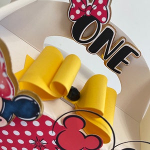 May include: A white cardboard box with a red and white polka dot design featuring a yellow bow, a black and red Mickey Mouse head, and the word "ONE" in black with a gold outline.