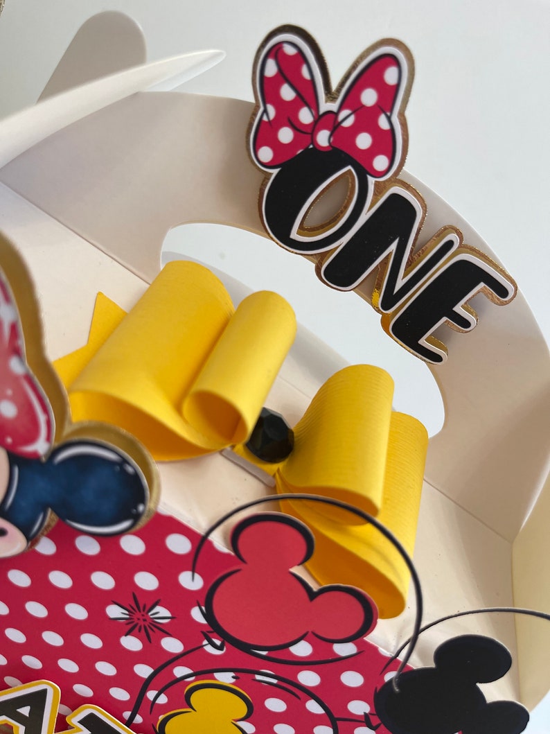May include: A white cardboard box with a red and white polka dot design featuring a yellow bow, a black and red Mickey Mouse head, and the word "ONE" in black with a gold outline.