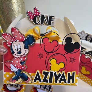 May include: A red and yellow Mickey Mouse themed party favor box with a Minnie Mouse character on the front. The box says "Aziyah" and "One" on it.