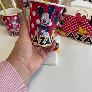 May include: Red and white paper cup with a Mickey Mouse design and the name "AZAI" printed on the bottom. The cup has white polka dots and a yellow straw.