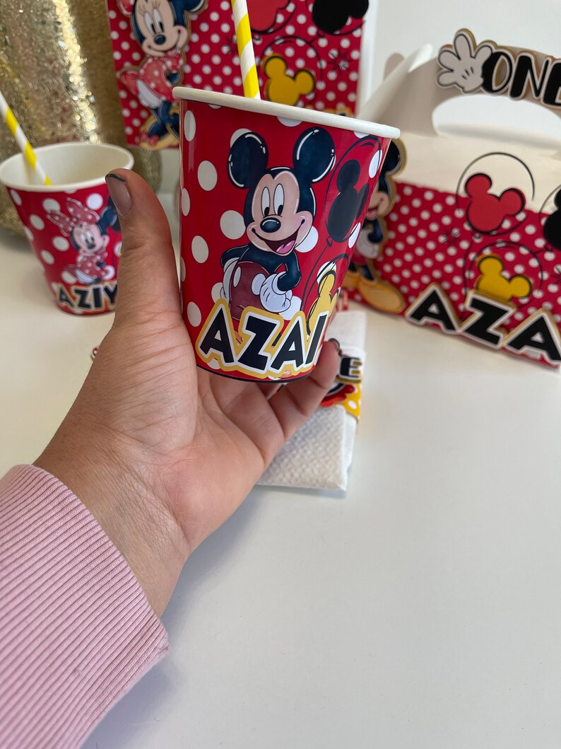 May include: Red and white paper cup with a Mickey Mouse design and the name "AZAI" printed on the bottom. The cup has white polka dots and a yellow straw.