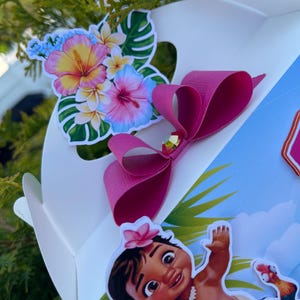 May include: A white gift box decorated with a pink bow and tropical flower stickers. The box features a cartoon image of a baby girl with a flower in her hair and a rooster. The flowers are pink, yellow, and blue.