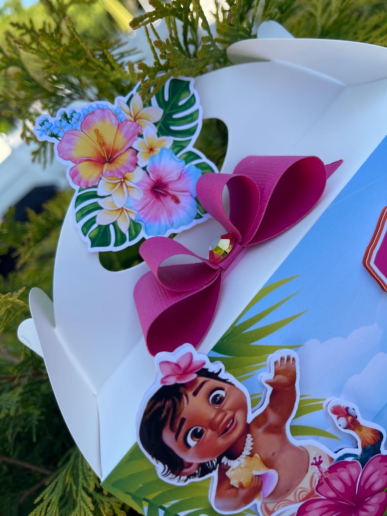 May include: A white gift box decorated with a pink bow and tropical flower stickers. The box features a cartoon image of a baby girl with a flower in her hair and a rooster. The flowers are pink, yellow, and blue.