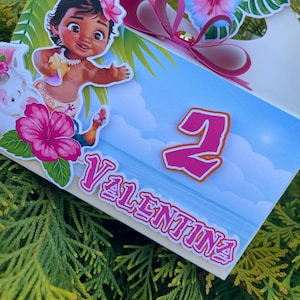 May include: A white gift box decorated with a cartoon image of a young girl, flowers, and the name "Valentina". The box features a pink bow and the number "2". The background is a blue sky with clouds.
