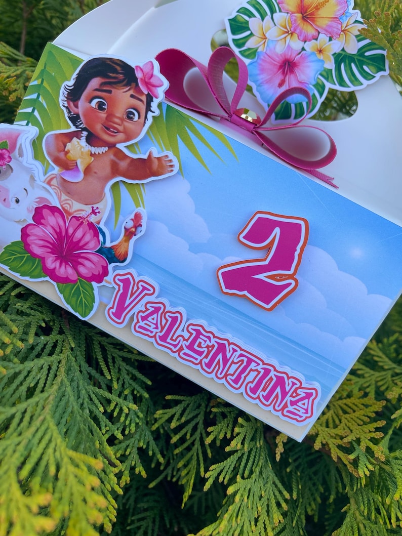 May include: A white gift box decorated with a cartoon image of a young girl, flowers, and the name "Valentina". The box features a pink bow and the number "2". The background is a blue sky with clouds.