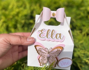 Puede incluir: Una caja de regalo blanca con un lazo rosa, una mariposa rosa y dorada y el nombre "Ella" en letras doradas. La caja también tiene un círculo rosa con la palabra "uno" en letras doradas.