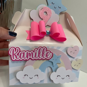 Puede incluir: Una caja de regalo de fiesta rosa y azul con un arcoíris, nubes y estrellas. La caja está decorada con el nombre "Kamille" y el número "9".