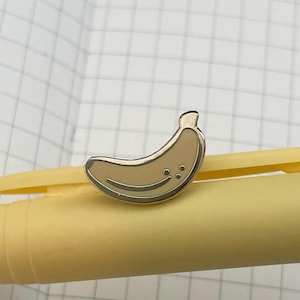 May include: A gold-colored banana enamel pin with a smiling face, resting on a yellow pen. The pen has a gray cap with the brand name "BIC" printed on it. The background is a white notebook with a grid pattern.