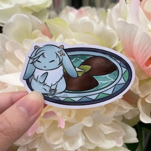 May include: A sticker featuring a light blue cartoon bunny with a cross on its forehead, sitting in a blue and white bowl with two brown chocolate-like treats. The bowl has a decorative border.