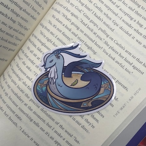 May include: A sticker featuring a blue and white illustration of a mythical creature with a long, serpentine body and a horned head. The creature is depicted in a bowl of soup with a green leaf floating on top. The sticker is on a page of a book with text about accepting a hug.