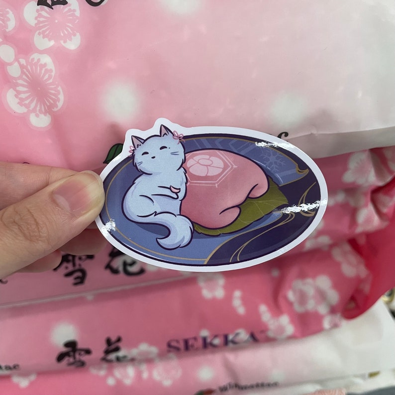 May include: A sticker featuring a cartoon white cat with pink accents, sleeping on a pink mochi with a flower design. The sticker is on a pink and white background with Japanese characters.