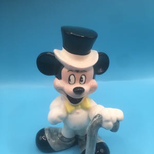 May include: A ceramic figurine of Mickey Mouse dressed in a tuxedo with a top hat and holding a cane. He is standing on a blue background.