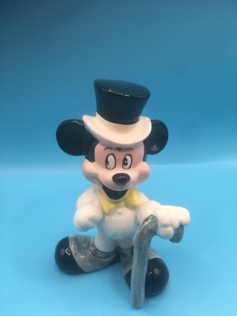 May include: A ceramic figurine of Mickey Mouse dressed in a tuxedo with a top hat and holding a cane. He is standing on a blue background.