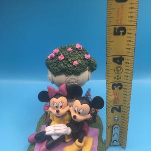 Puede incluir: Una figurita de cer&aacute;mica de Mickey Mouse y Minnie Mouse sentados en una manta de picnic con un arbusto de flores rosas detr&aacute;s. La figurita mide aproximadamente 13 cm de alto.