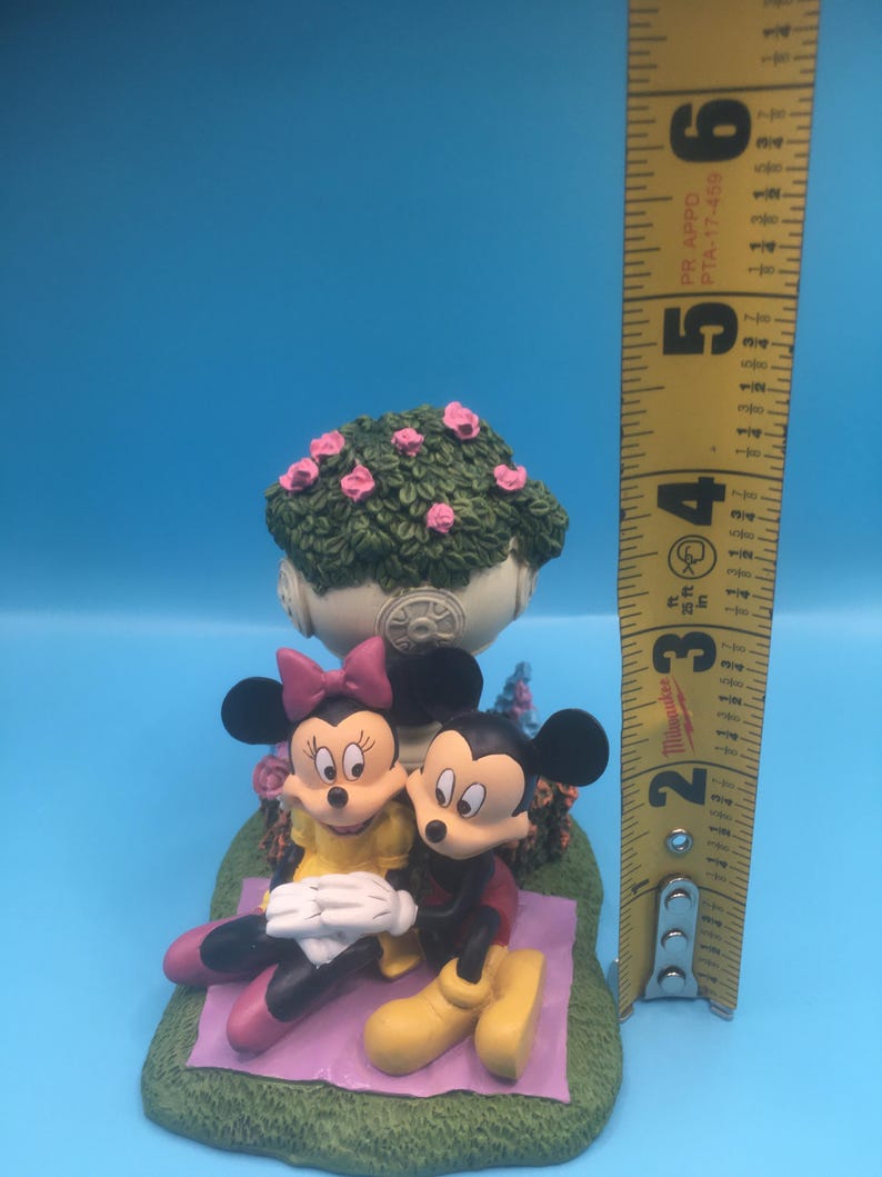 Puede incluir: Una figurita de cer&aacute;mica de Mickey Mouse y Minnie Mouse sentados en una manta de picnic con un arbusto de flores rosas detr&aacute;s. La figurita mide aproximadamente 13 cm de alto.