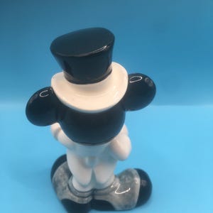 May include: A ceramic figurine of a cartoon character wearing a black top hat and a white tuxedo. The figurine is standing with its hands clasped in front of it. The figurine is painted in black, white, and gray.