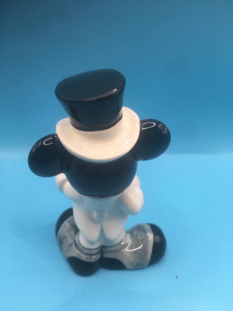 May include: A ceramic figurine of a cartoon character wearing a black top hat and a white tuxedo. The figurine is standing with its hands clasped in front of it. The figurine is painted in black, white, and gray.