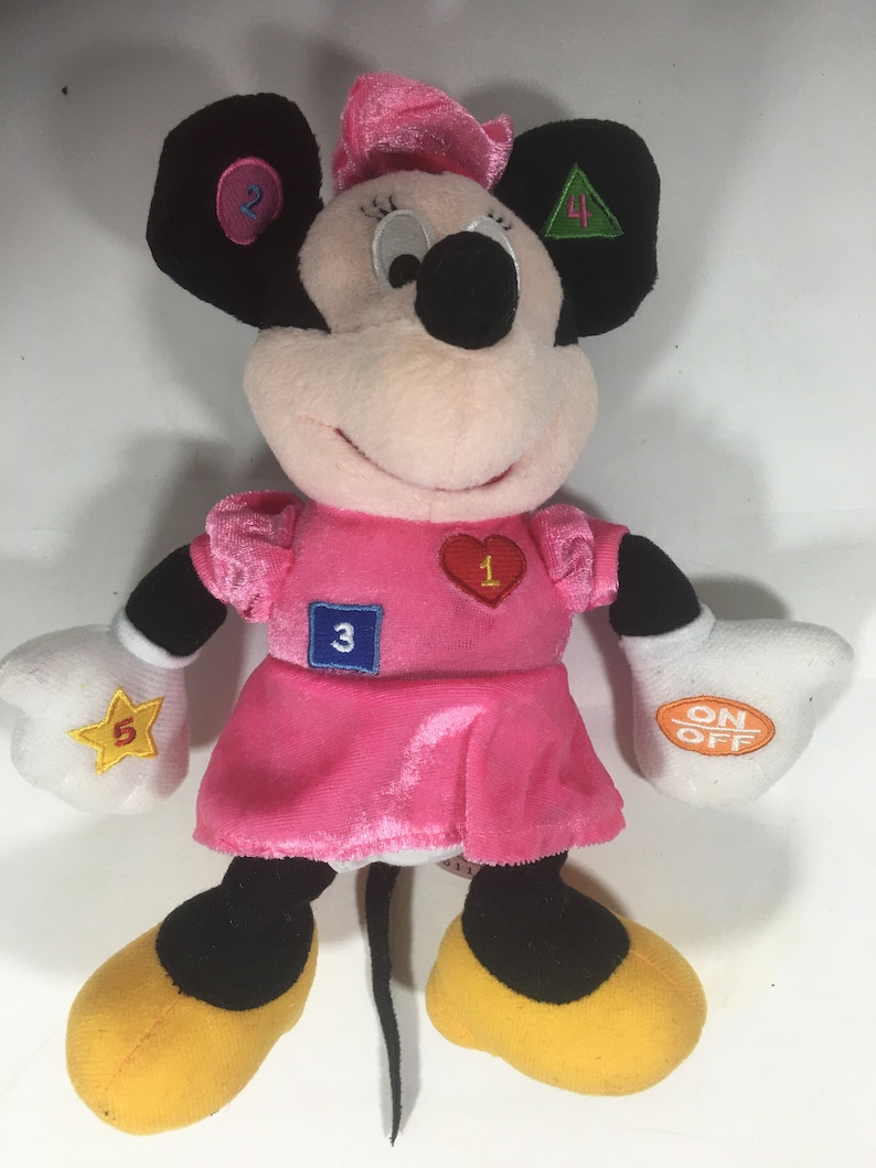 May include: A pink plush Minnie Mouse toy with a pink dress and yellow shoes. The toy has embroidered numbers 1 through 5 on its dress and ears. The toy has an on/off switch on its right hand.