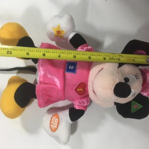 May include: Pink and black plush toy of a cartoon mouse wearing a pink dress and a white shirt with numbers 1, 3, and 5 on it. The toy has a black tail and yellow shoes. The toy has an on/off switch on its hand.