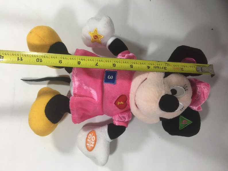 May include: Pink and black plush toy of a cartoon mouse wearing a pink dress and a white shirt with numbers 1, 3, and 5 on it. The toy has a black tail and yellow shoes. The toy has an on/off switch on its hand.