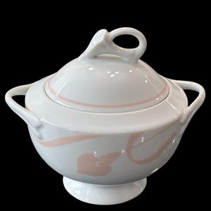 May include: White ceramic sugar bowl with a lid and two handles. The bowl features a pink floral design and a pink line around the lid. The lid has a loop handle. The bowl sits on a small pedestal base.
