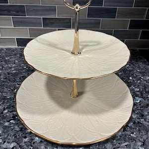 Può includere: Un vassoio a due livelli con manico e finiture dorate. I piatti bianco sporco hanno un motivo strutturato a forma di foglia. Il vassoio è progettato per esporre e servire cibo.