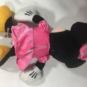 May include: Pink and black plush toy of a cartoon mouse wearing a pink dress. The toy has a yellow nose and large yellow feet.