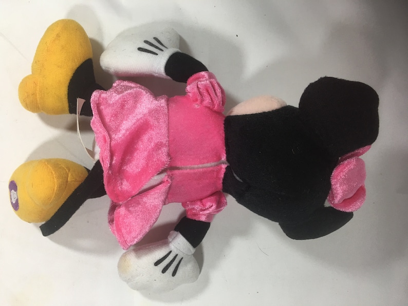 May include: Pink and black plush toy of a cartoon mouse wearing a pink dress. The toy has a yellow nose and large yellow feet.