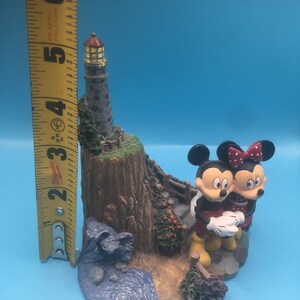 以下が含まれることがあります： 岩場の海岸に建つ灯台のそばに、ミッキーマウスとミニーマウスの陶器製フィギュアが立っています。灯台は茶色と灰色で、赤い屋根と黄色い窓が付いています。フィギュアは明るい色で塗られており、幻想的なスタイルです。