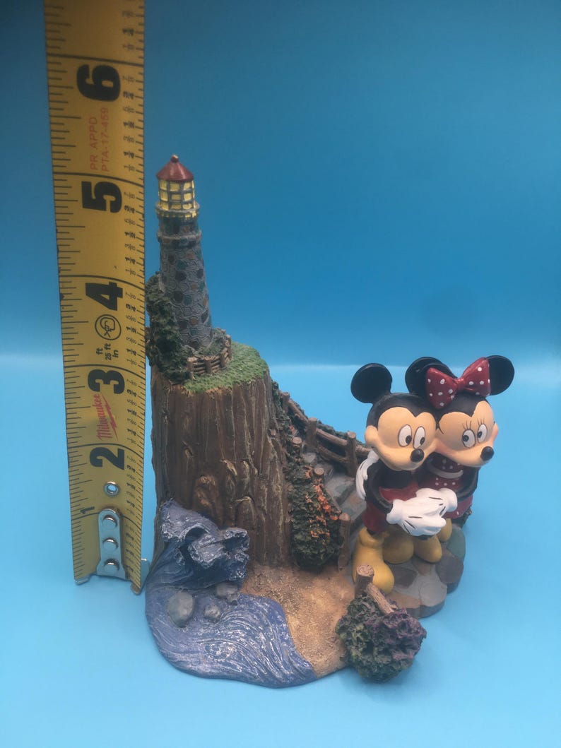 以下が含まれることがあります： 岩場の海岸に建つ灯台のそばに、ミッキーマウスとミニーマウスの陶器製フィギュアが立っています。灯台は茶色と灰色で、赤い屋根と黄色い窓が付いています。フィギュアは明るい色で塗られており、幻想的なスタイルです。