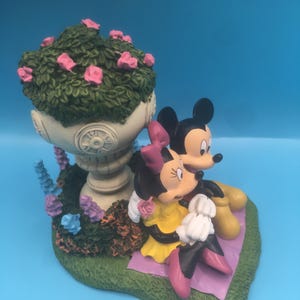 Puede incluir: Una figurita de cer&aacute;mica de Mickey Mouse y Minnie Mouse sentados en una manta de picnic. Mickey lleva pantalones cortos rojos y zapatos amarillos. Minnie lleva un vestido amarillo y zapatos rosas. Est&aacute;n sentados frente a una maceta blanca con hojas verdes y flores rosas.