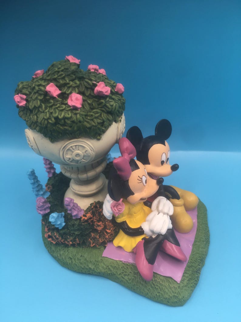 Puede incluir: Una figurita de cer&aacute;mica de Mickey Mouse y Minnie Mouse sentados en una manta de picnic. Mickey lleva pantalones cortos rojos y zapatos amarillos. Minnie lleva un vestido amarillo y zapatos rosas. Est&aacute;n sentados frente a una maceta blanca con hojas verdes y flores rosas.