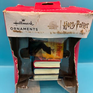 Może przedstawiać: Czerwono-białe pudełko Hallmark Ornaments z marką Harry Potter. Wewnątrz widoczne są miniaturowe książki i figurka. Pudełko jest otwarte, ukazując zawartość. Widoczne są również słowa "WIZARDING WORLD".