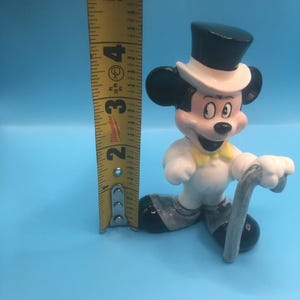 May include: A white ceramic figurine of Mickey Mouse wearing a black top hat, white bow tie, and a white tuxedo. He is holding a silver cane and is standing on a blue surface. The figurine is approximately 4 inches tall.