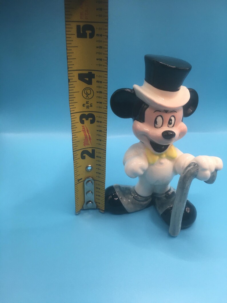 May include: A white ceramic figurine of Mickey Mouse wearing a black top hat, white bow tie, and a white tuxedo. He is holding a silver cane and is standing on a blue surface. The figurine is approximately 4 inches tall.
