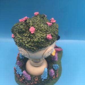 Puede incluir: Una urna de jard&iacute;n de cer&aacute;mica blanca con una parte superior verde frondosa y flores rosas. La urna est&aacute; rodeada de un lecho de flores moradas y azules.