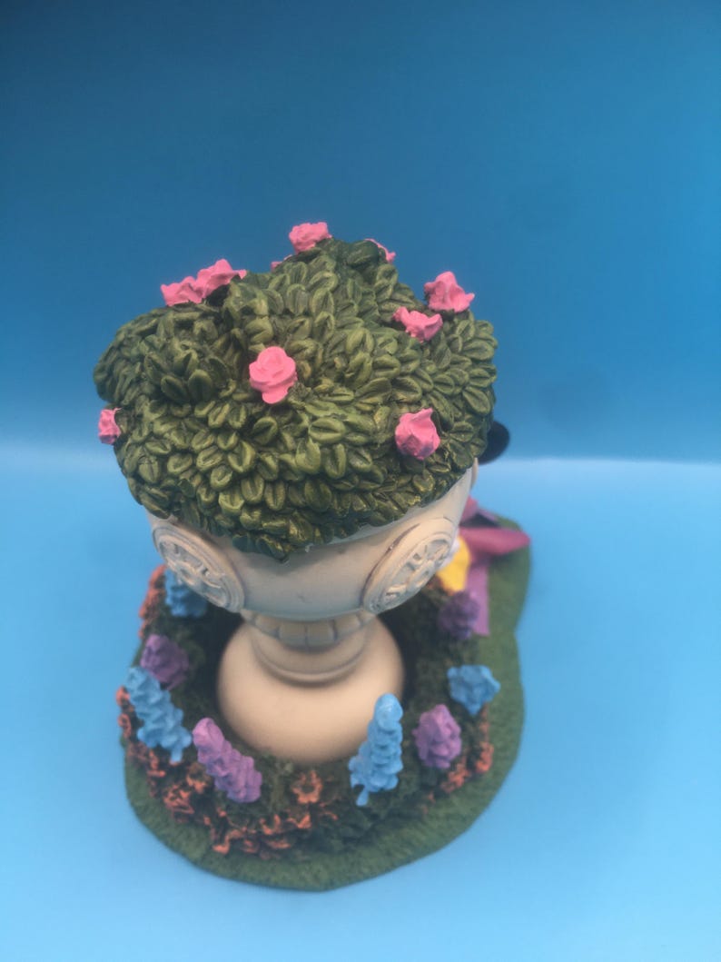 Puede incluir: Una urna de jard&iacute;n de cer&aacute;mica blanca con una parte superior verde frondosa y flores rosas. La urna est&aacute; rodeada de un lecho de flores moradas y azules.