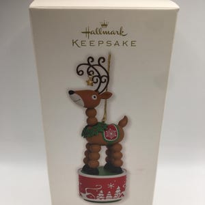 以下が含まれることがあります： ホールマーク・キープセイクのクリスマスオーナメント。首に緑のリースを巻いた茶色と赤のゆらゆら揺れるトナカイ。オーナメントは雪の結晶と冬の風景が描かれた赤と白の台座の上に置かれています。
