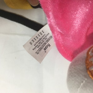 May include: A white tag with black text that reads "Kcare KIU HUNG INDUSTRIES LTD. 63 Au Pui Wan St., Fotan, Hong Kong. 14/F, Yale Ind. Centre, E-mail: kh@kluhung.com 15111K H" attached to a pink and white plush toy.