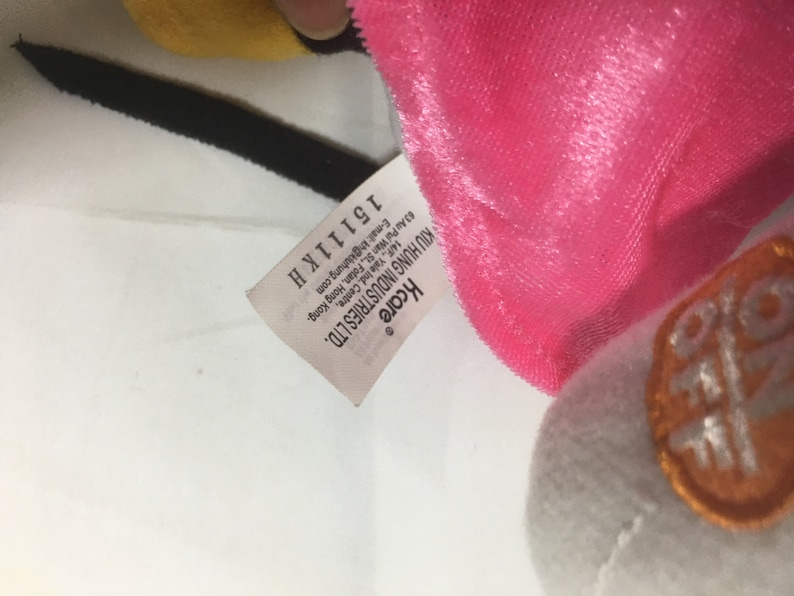 May include: A white tag with black text that reads "Kcare KIU HUNG INDUSTRIES LTD. 63 Au Pui Wan St., Fotan, Hong Kong. 14/F, Yale Ind. Centre, E-mail: kh@kluhung.com 15111K H" attached to a pink and white plush toy.