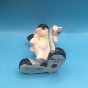 May include: A white ceramic figurine of Mickey Mouse dressed in a tuxedo, holding a cane. The figurine is standing on a black base with the words "COISNEY JAPAN" printed on it.