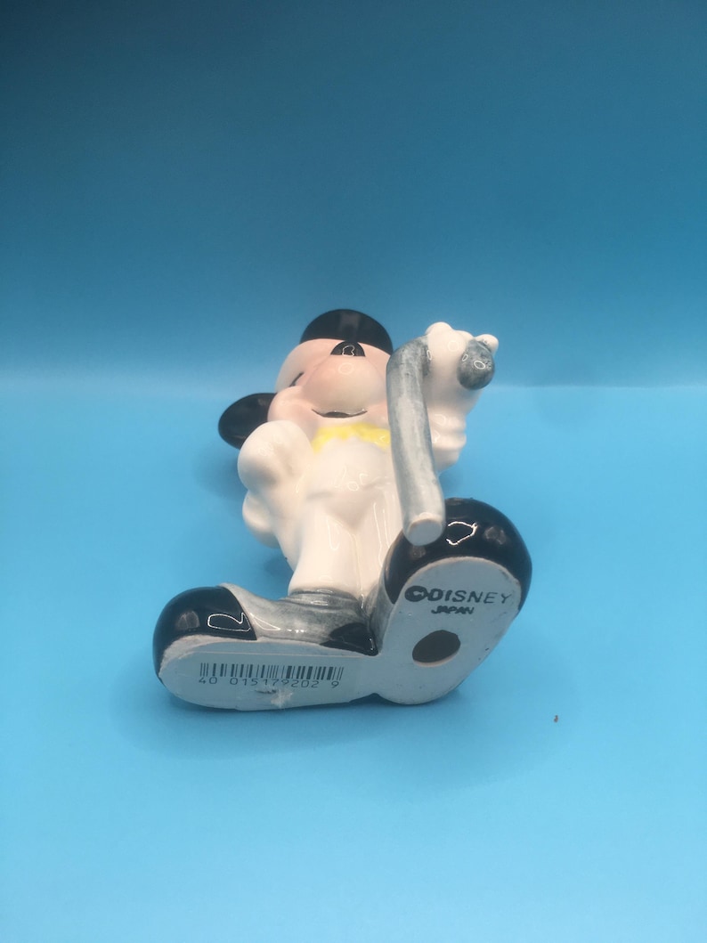 May include: A white ceramic figurine of Mickey Mouse dressed in a tuxedo, holding a cane. The figurine is standing on a black base with the words "COISNEY JAPAN" printed on it.