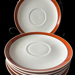 May include: Set of six white ceramic saucers with a wide orange band around the edge. The saucers are stacked on top of each other.