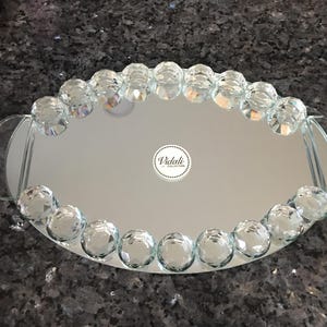 May include: Oval-shaped mirrored tray with a clear glass beaded border. The tray has two handles on the sides. The mirror has a reflective surface and the text "Vidali Collection" is visible in the centre.