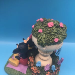 Puede incluir: Una figurita de cer&aacute;mica de Mickey Mouse y Minnie Mouse sentados en una manta en un entorno de jard&iacute;n. La figurita presenta una maceta blanca con follaje verde y flores rosas, rodeada de flores moradas y azules.