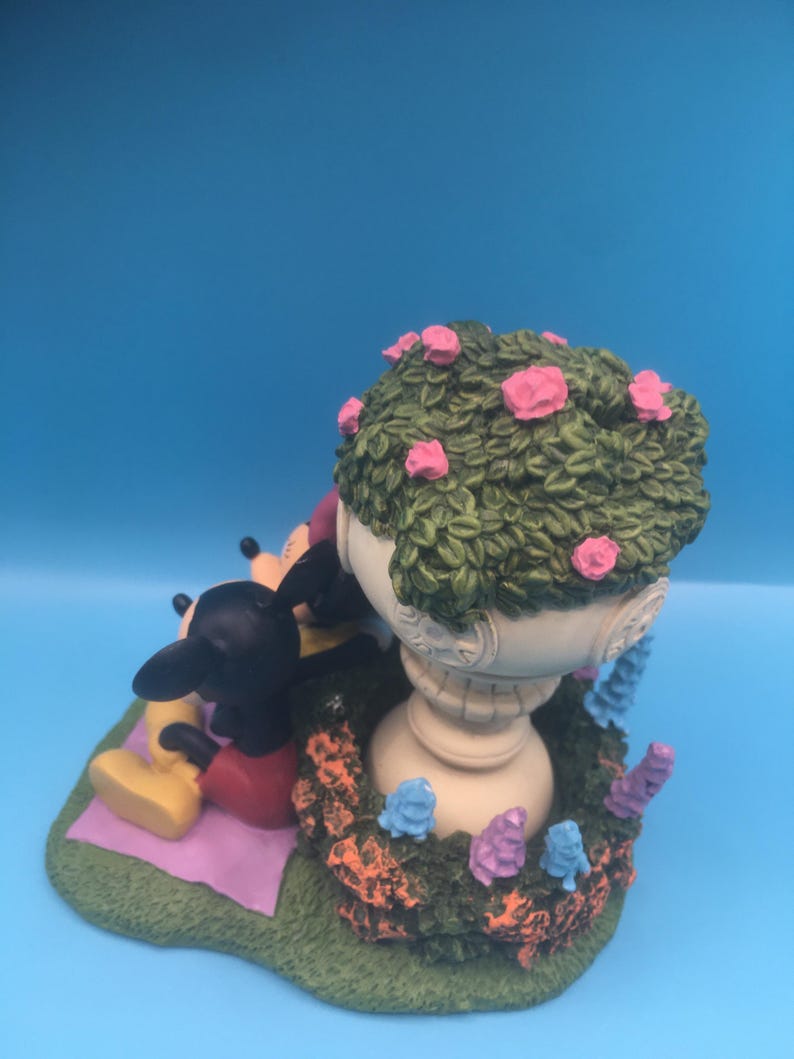 Puede incluir: Una figurita de cer&aacute;mica de Mickey Mouse y Minnie Mouse sentados en una manta en un entorno de jard&iacute;n. La figurita presenta una maceta blanca con follaje verde y flores rosas, rodeada de flores moradas y azules.