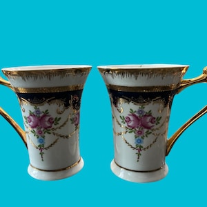 May include: Two white porcelain teacups with gold trim and floral designs. The cups have blue accents and gold handles.