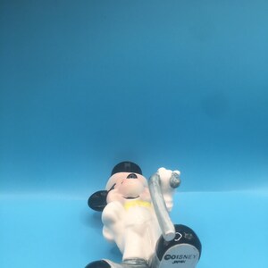 May include: A white ceramic figurine of Mickey Mouse dressed as a conductor, holding a baton. He is wearing a black top hat, a white shirt with a yellow bow tie, and black shoes. The figurine is made in Japan and has the Disney logo on the bottom.